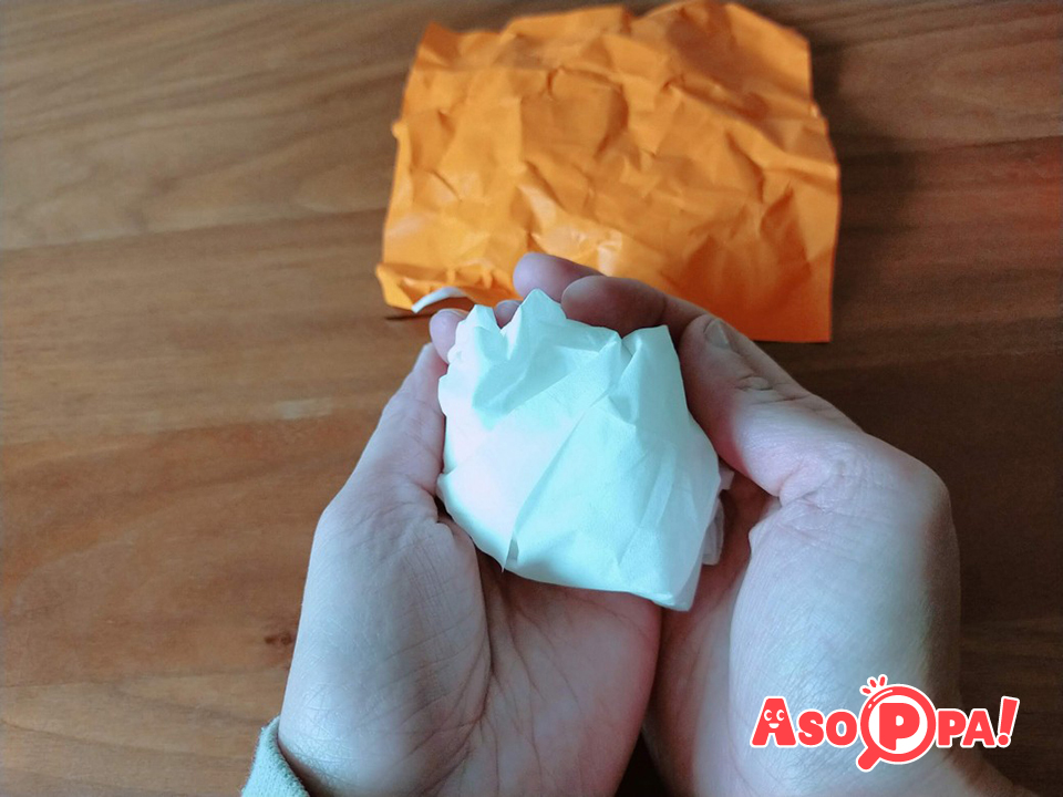 次にみかんを作ります。だいだい色の折り紙をくしゃっと丸めて広げ、シワを付けます。その折り紙で丸めたティッシュペーパー（５～６枚）を包み、テープでとめます。